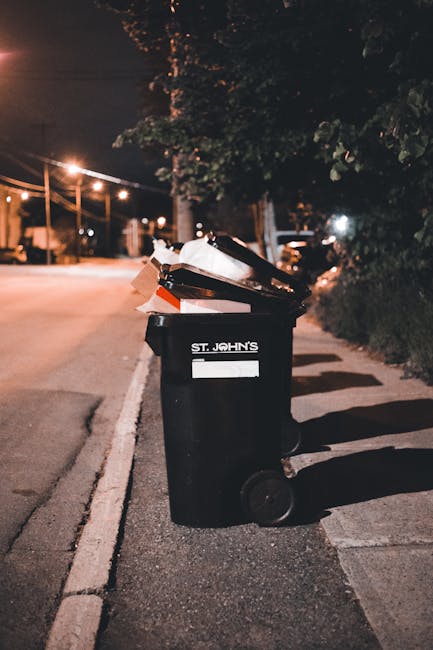 A black wheeled refuse bin positioned on the edge of a paved street at night, marked with the label 'St. John's', filled with various types of waste such as cardboard, plastic containers, and paper. T
